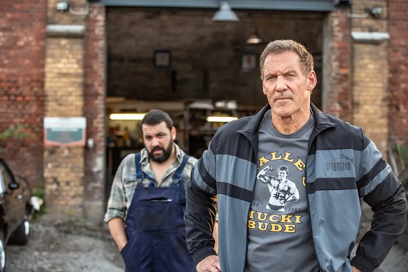 Characters from Der letzte Bulle stand outside a brick - building, one in a Puma jacket and the other in overalls.